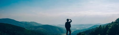 A lone traveler stands on a hilltop, gazing over a vast mountain landscape under a clear blue sky.